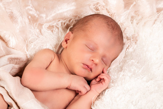 A baby boy is peacefully sleeping during his first professional photo shoot. He is dressed in white and covered with a white blanke
