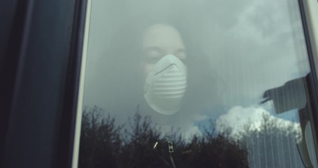 pensive young woman wears a mask to protect herself from covid-19 while looking out the window.  a bird in flight is reflected on the window glass - Powered by Adobe