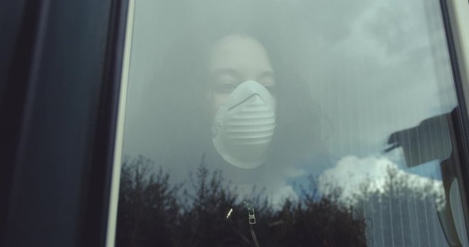 pensive young woman wears a mask to protect herself from covid-19 while looking out the window.  a bird in flight is reflected on the window glass