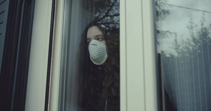 pensive young woman wears a mask to protect herself from covid-19 while looking out the window