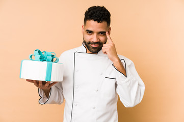 Young latin baker man holding a cake isolated pointing temple with finger, thinking, focused on a task.