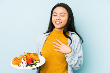 Young chinese woman holding waffle isolated on blue background  smiling and raising thumb up