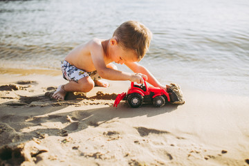 Caucasian child boy plays toy red tractor, excavator on sandy beach by the river in shorts at sunset day
