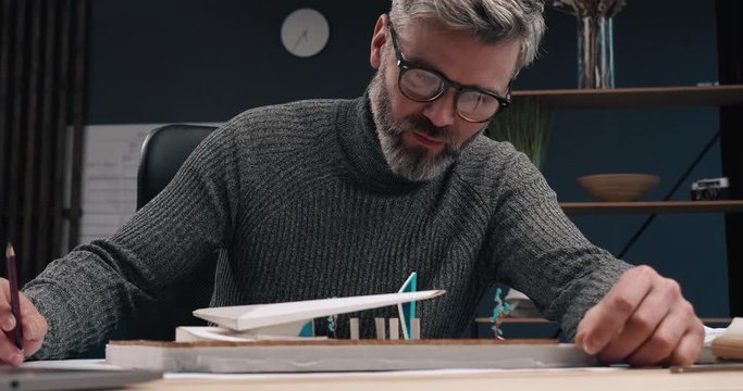 Serious and Successful Architect Looking at his Model of Futuristic Building with park near it. Looking satisfied with his Job. Having Eyeglasses and Gray Beard. Handsome man of Middle Age.