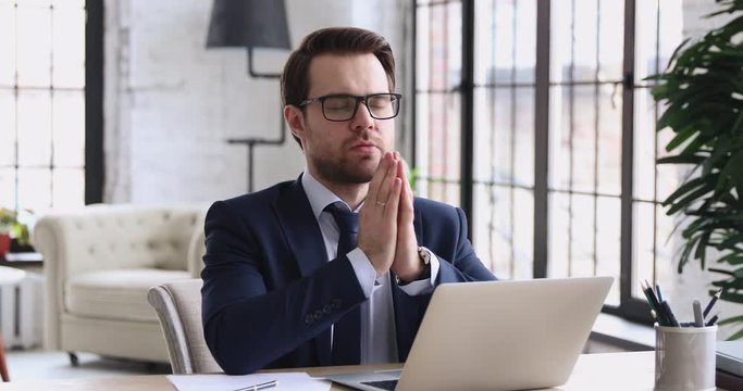 Serious focused businessman wearing suit praying at workplace with eyes closed. Faithful male executive saying prayer or meditating with namaste put hands together feels nervous sitting at work desk.
