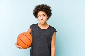 Poster Kid boy playing basketball isolated on blue background confused, feels doubtful and unsure. © Asier