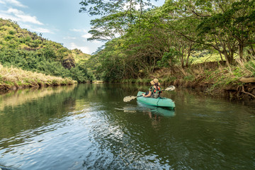 Fototapeta na wymiar Girl, woman, right, paddling a green kayak on a calm river with trees line the bank and puffy clouds in the sky, Wailua River, Kauai, Hawaii