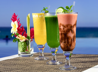 Three colorful smoothies and tropical flowers on pool and sea background