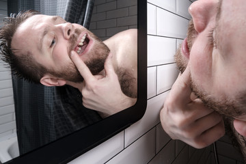 Man opened his mouth and carefully examines the absence of a tooth in mirror, peering out from behind the curtains in the shower.