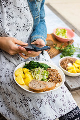Woman take picture of food with phone at kitchen or summer cafe. Smartphone photography of vegetables dinner or lunch for blogging or social media publications. Vegan vegetarian healthy diet.