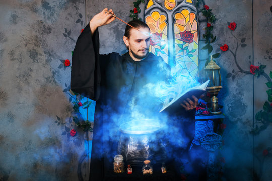 The mage is cooking a potion. A male wizard in a black mantle with a magic wand and a book. He casts a spell over the witch's cauldron. Guy in an old castle with luminous magic
