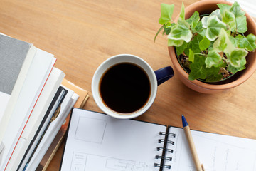 A cup of coffee next to a pile books and a notebook. A pot of ivy gives a refreshing look to the scene. Elements give a clue to break time after hard work.