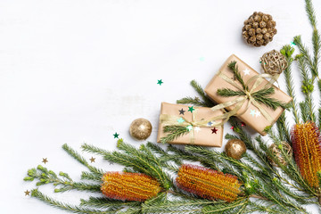 Australian native plant Banksia frames the Christmas inspired background. Gifts, gold ribbon, glitter, cream pine cones and other Christmas decorations on a rustic white background.
