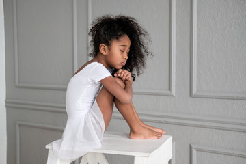 A portrait of an African ballerina in a white gymnastic costume sits on a wooden chair. © evelinphoto