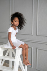 A portrait of an African ballerina in a white gymnastic costume sits on a wooden chair.