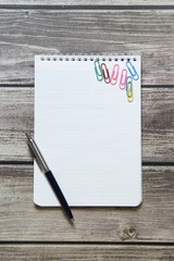 Notepad with a blank white sheet in a checker paper and with ball pen and with paper clips lies on the background of wooden boards