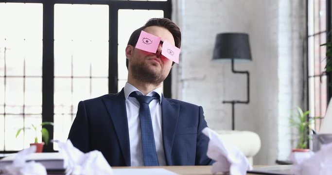 Funny exhausted businessman sleeping at workplace with drawn eyes on sticky notes on face. Overworked lazy male executive wearing suit napping sitting at work desk. Lack of sleep in office concept.