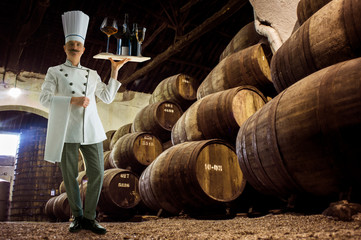the sommelier stands in the basement next to barrels of wine and holds a variety of alcohol on a...