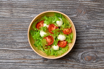 Fresh salad with cherry tomatoes, mozzarella cheese, greens leaves in bowl. Healthy summer salad vegetarian meal concept. Tomatoes and lettuce home made caprese salad on wood, top view