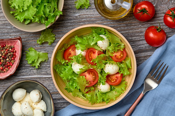 Fresh salad with cherry tomatoes, mozzarella cheese, greens leaves in bowl. Healthy summer salad vegetarian meal concept. Tomatoes and lettuce home made caprese salad on wood, top view