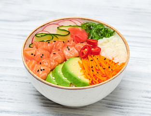 Hawaii salmon poke, avocado, rice, seaweed, cucumber, radish, carrot in bowl. Home made vegetarian poke. Seafood concept. Asian salmon poke salad on white wood background.