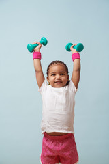 Portrait of cute little girl lifting small colored dumbbells overhead against pale blue background....