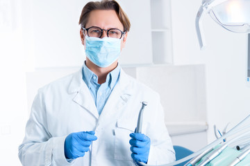Man in white doctor robe uniform, glasses, mask, gloves is holding instrument, standing in office, clinic. Dentist workplace. Orthodontist is preparing for visit of patient. Dentistry concept.