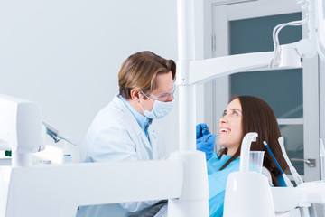 Smiling young woman is sitting in dental blue chair in clinic, office. Man doctor, orthodontist is...