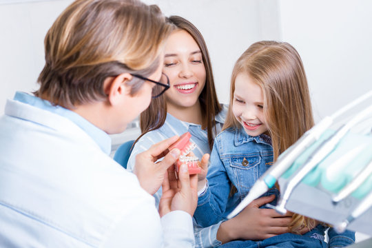 Little Cute Smiling Girl Is Sitting In Dental Chair With Mother In Clinic, Office. Doctor Is Preparing For Examination Of Child Teeth, Talking With Patient. Visiting Dentist With Children.