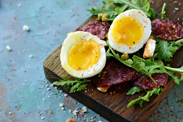 Soft-boiled eggs for Breakfast with chorizo ​​sausage, fresh arugula salad and croutons (crackers) on a wooden board on a blue background.