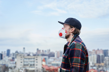 Young man in hat with respirator standing in front of the city, coronavirus disease and world...