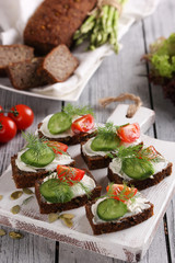 Healthy food. Sliced dark bread with cream cheese, cucumber, red cherry tomatoes and dill on a white wooden board. Pumpkin, flax seeds, lettuce, asparagus on a light wooden background. Copy space