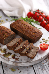 Fototapeta na wymiar Healthy food. Sliced dark bread with pumpkin seeds and flax seeds on a white wooden board. Red cherry tomatoes, arugula on a light wooden background. Background image, copy space