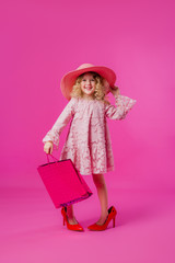 little girl in her mother's big shoes, dress, hat, and shopping bags. baby girl smiling and holding bags from the store. little fashionista on a shopping trip