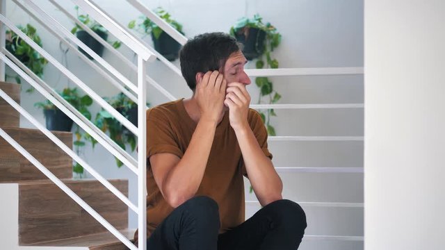 Pensive sad and depressed young man sitting alone on stairs at home: painful sad man.
