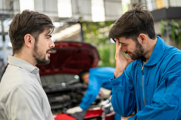 The mechanic in the blue jumpsuit is checking the engine in front of the red open car trunk and the man who is the  car’s owner is talking with the checker who putting the right hand at his forehead