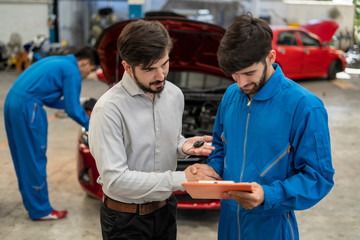 The mechanic in the blue jumpsuit is checking the engine in front of the red open car trunk and the man who is the  car’s owner is talking with the checker who putting the right hand at his forehead