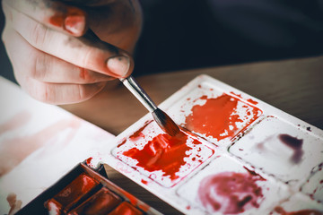 The artist's dirty hand holds a paintbrush with which she mixes scarlet watercolor paint on a...