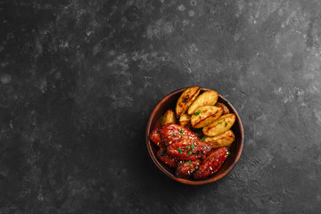Grilled chicken wings in barbecue sauce with sesame and parsley, with rustic potatoes in a wooden plate on a dark table. Top view with space for text. Delicious beer appetizer on a dark background.
