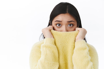 Close-up portrait timid and insecure cute asian girl pull sweater collar on face as feel afraid and scared of horror movie, eyes exress fear, trembling frightened, standing white background - obrazy, fototapety, plakaty