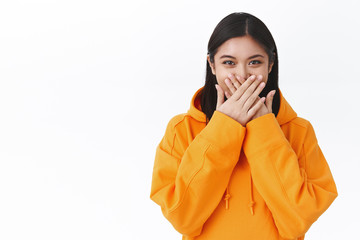 Waist-up portrait funny and cute asian girl in orange hoodie, giggle hiding laugh, trying calm, smiling with eyes while press hands to lips, cover mouth palms, standing white background
