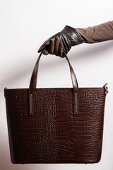 girl's hand in gloves holds brown leather bag on light background, leather bag in hand side view on light background, leather handbag holds female hand on light background, crocodile leather bag
