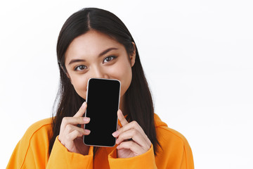 Close-up portrait of cute asian girl in orange hoodie, hiding face behind smartphone, showing mobile phone display, smiling with eyes, recommend application, app promo, white background