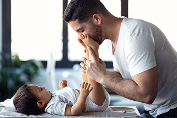 Smiling young father has fun with little baby while changing his nappy at home.