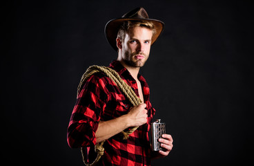 Fototapeta premium Brutal cowboy drinking alcohol. Western culture. Man handsome unshaven cowboy black background. Western life. Man wearing hat hold rope and flask. Lasso tool of American cowboy. Sheriff concept