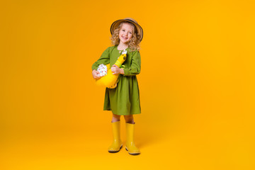 little girl smiles in a green linen dress, yellow rubber boots and a straw hat holding a textile toy chicken on a yellow background of isolate. concept of eco-friendly food and farming