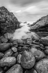A vertical black and white sunset landscape photograph of misty waves crashing on the rocks by the South Coast in South Africa