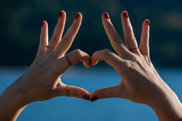 Female hands in heart shape in focus, blue sea and beachfront on background 