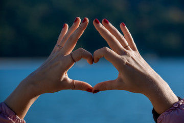 Female hands in heart shape in focus, blue sea and beachfront on background 