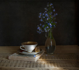 Still life with a cup of coffee with a saucer on an old book on a wooden table, near a glass vase with small blue flowers, dark background © Astibuk